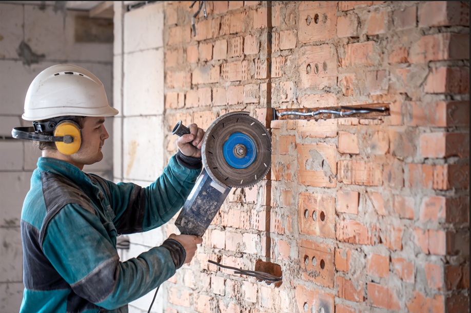 A construction worker wearing a hard hat and ear protection uses a wall chaser to cut precise grooves into a brick wall for electrical conduit installation.
