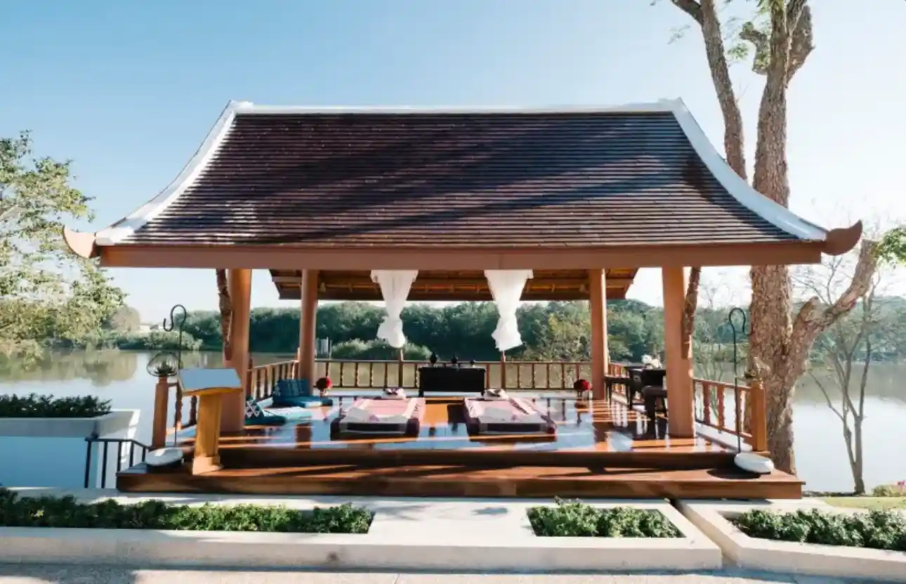 An elegant open-air pergola with a traditional tiered roof overlooks a serene lake, featuring an interior arranged with floor mats and white linens for a peaceful lakeside massage or meditation session.