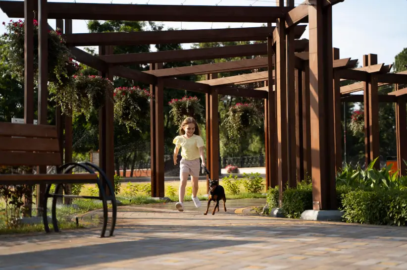 
Gemini said
A young girl happily runs along a paved park walkway while leading her small dog on a leash through a series of wooden garden pergolas adorned with hanging flower baskets.