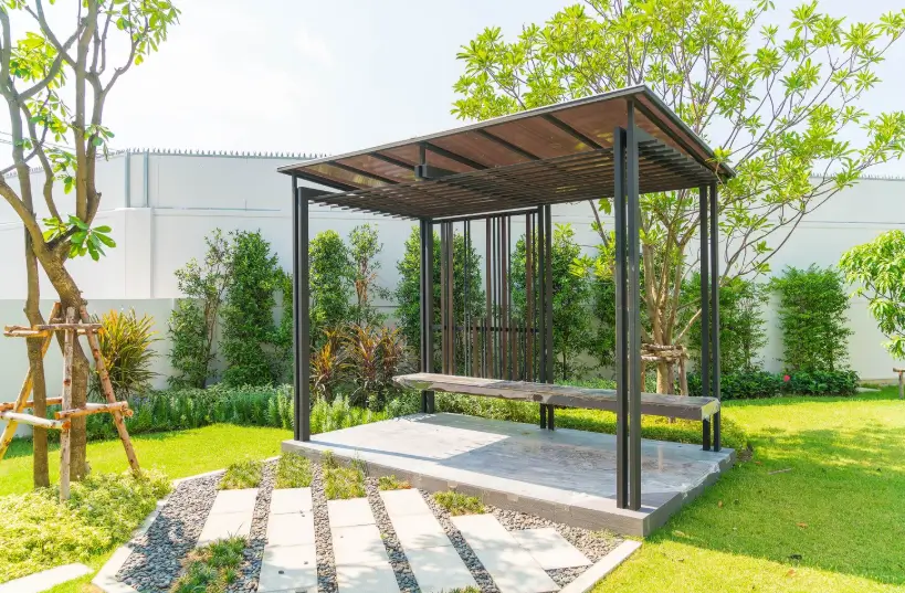 A modern, minimalist garden pergola with a dark metal frame and slatted roof provides a shaded seating area over a stone platform in a lushly landscaped backyard.