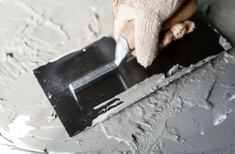 A gloved hand uses a metal trowel to smooth out wet cement, creating a level surface that will eventually be ready for a concrete sealer.