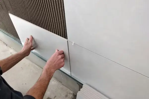 A person installs a large, white ceramic tile onto a bathroom remodel, wall layered with combed adhesive mortar.