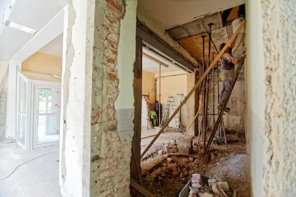 A worker stands on a ladder inside a building undergoing a major structural renovation with exposed brick, rubble, and temporary supports.