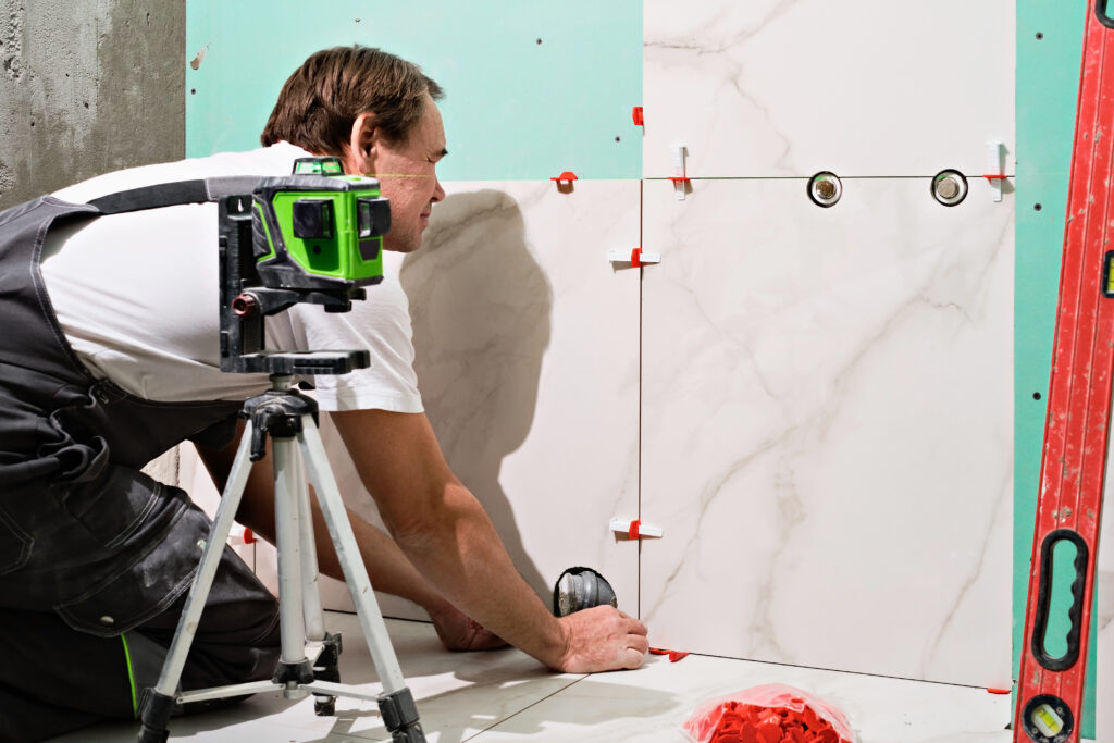 A professional construction worker in overalls is using a laser level and tile spacers to carefully install large-format marble-patterned porcelain tiles on a bathroom wall.