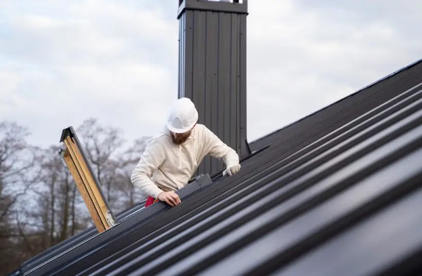 A professional contractor wearing a hard hat examines the flashing near a chimney during a comprehensive roof repair of a modern metal roofing system.