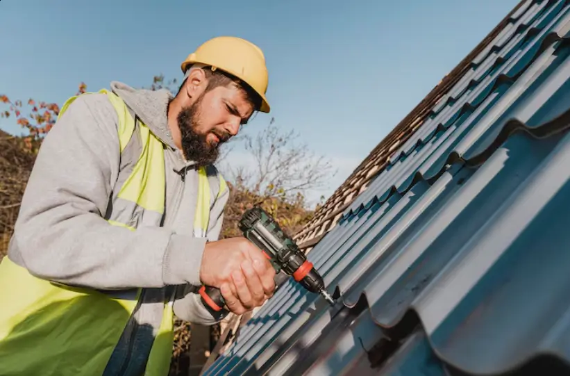 A construction worker in a hard hat and safety vest uses a power drill to secure metal panels, a vital step in a professional roof repair.