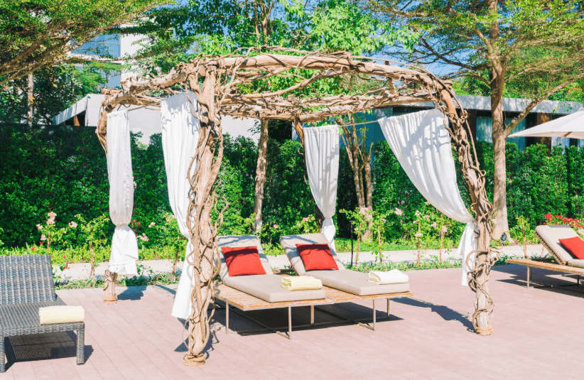 Sunlight filters through a rustic wooden structure draped with white fabric and climbing vines, creating an elegant pergola design over comfortable poolside lounge chairs.