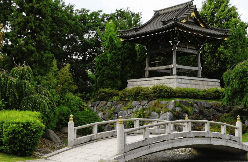 A traditional stone bridge leads toward an ornate wooden structure with a tiered roof nestled among lush greenery, showcasing a classic and cultural pergola style.