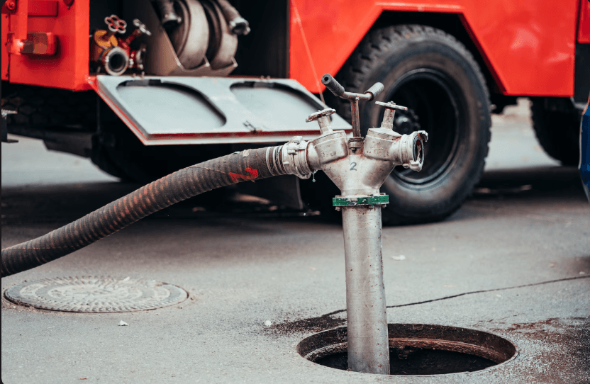 A metal standpipe connected to a heavy-duty hose emerges from an open manhole near a service vehicle, representing the specialized equipment used for advanced drainage systems.