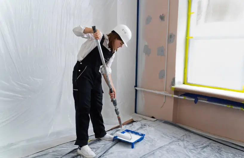 A painter in a white hard hat and black overalls uses an extension pole to load a roller with fresh white paint, a key step in providing professional painting services.
