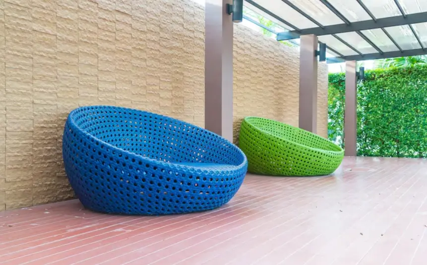 Two stylish, colorful wicker patio furniture chairs sit on a sleek wooden deck beneath a modern structure with translucent panels.