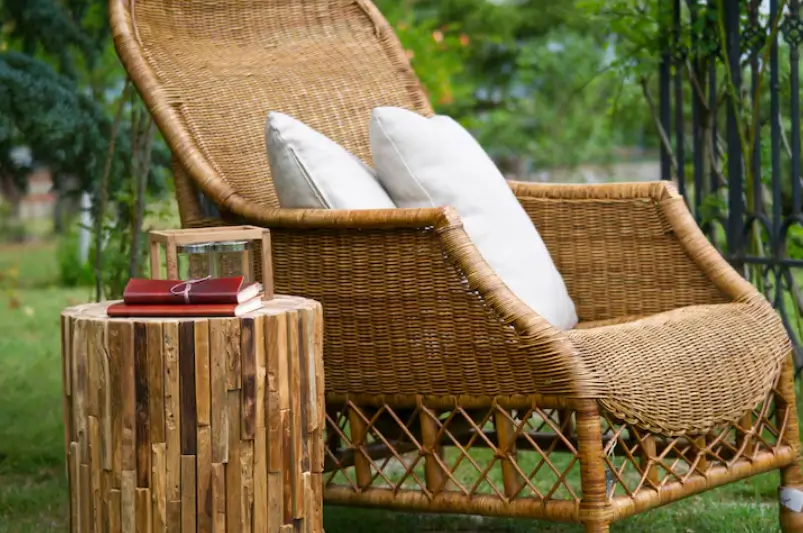 A comfortable wicker armchair with white cushions sits next to a rustic wooden side table in a lush garden, showcasing elegant patio furniture for outdoor relaxation.