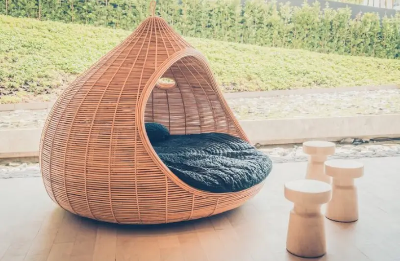A stylish teardrop-shaped wicker daybed with dark cushions sits on a modern deck alongside minimalist stools, offering a luxurious example of contemporary patio furniture.
