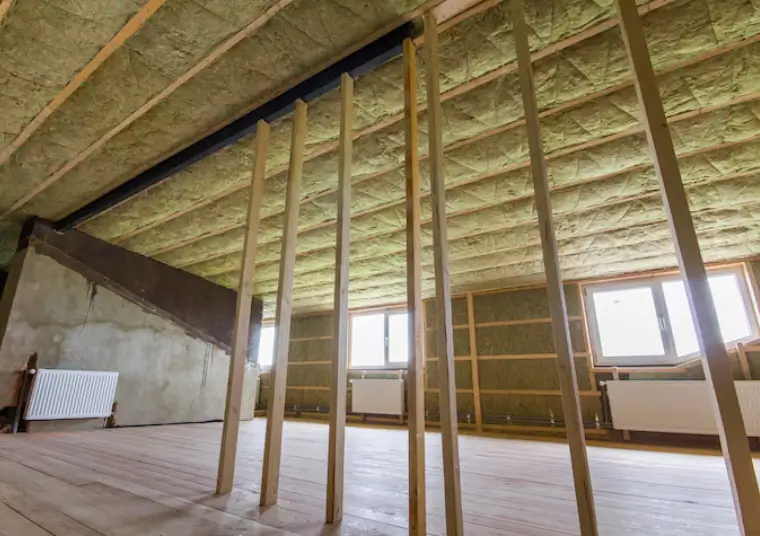 A spacious, unfinished attic features wooden framing studs alongside thick rolls of exposed green fiberglass lining the ceiling and walls, showcasing a common selection of insulation materials for residential temperature regulation.