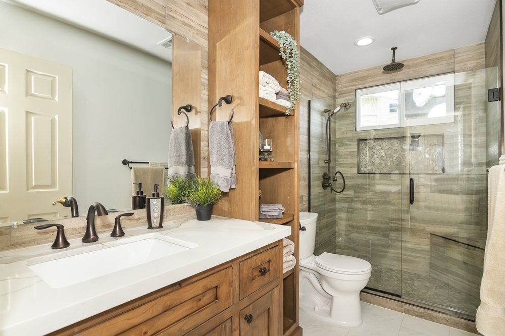 Bathroom remodel with a wood vanity, open shelving and a glass walk-in shower by Akraba Remodeling